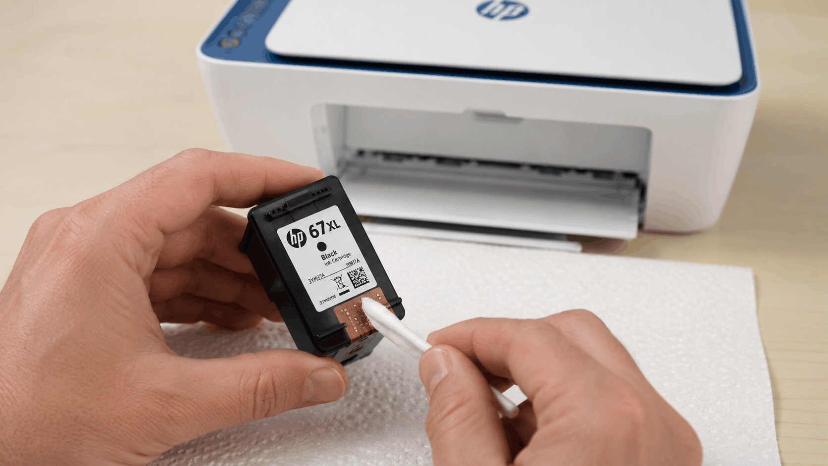 A close-up of someone gently wiping the copper nozzle plate on the underside of an HP 67 ink cartridge with a damp lint-free cloth.